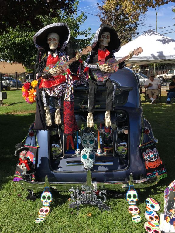 Two dressed skeletons playing painted guitars sit on top of an open truck filled with candles and stylized paper skulls