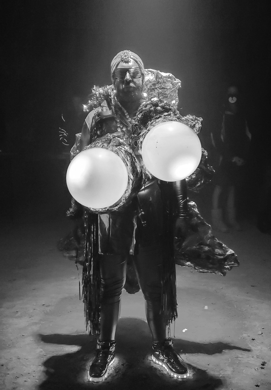 Black and white photograph of a person in an elaborate, shiny costume with two large glowing orbs