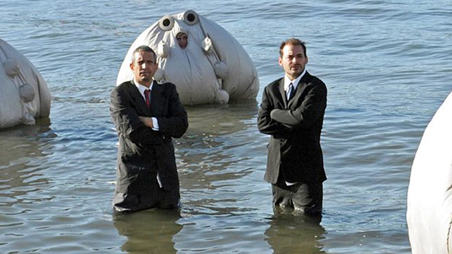 Photo of two men in suits sanding in knee-deep water with three figures in large inflated white fabric surrounding them.