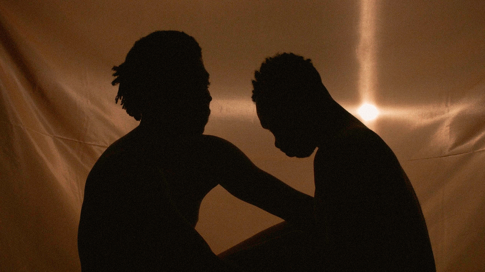 Image of two silhouettes sitting facing one another against a backlit gold cloth
