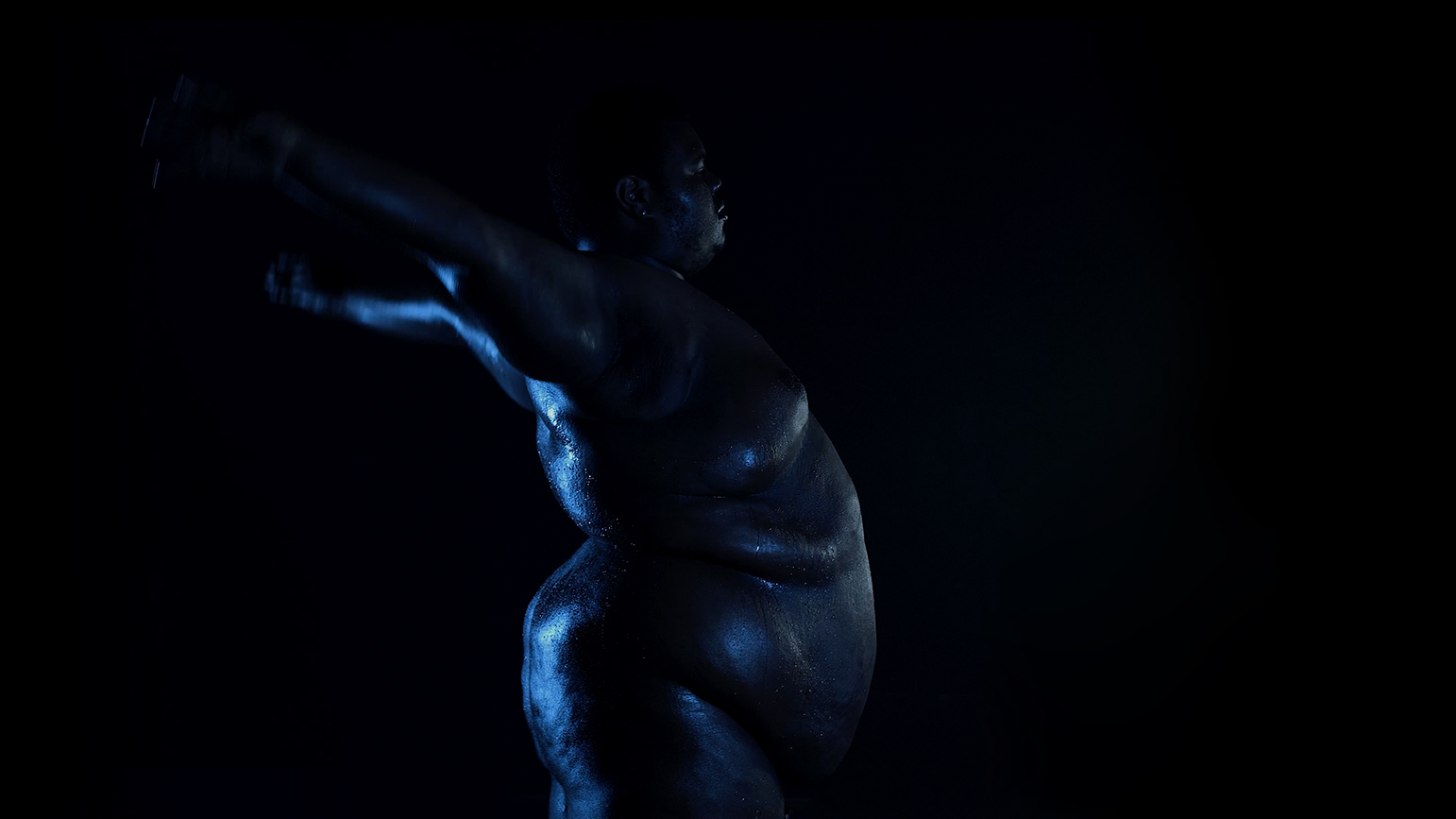 Video still of a nude figure in very low lighting from the side with arms reaching backward