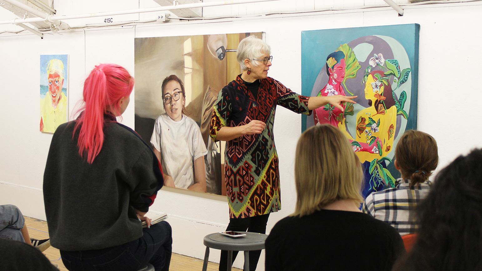 Professor standing in front of and pointing to a painting while students sit facing her and listening