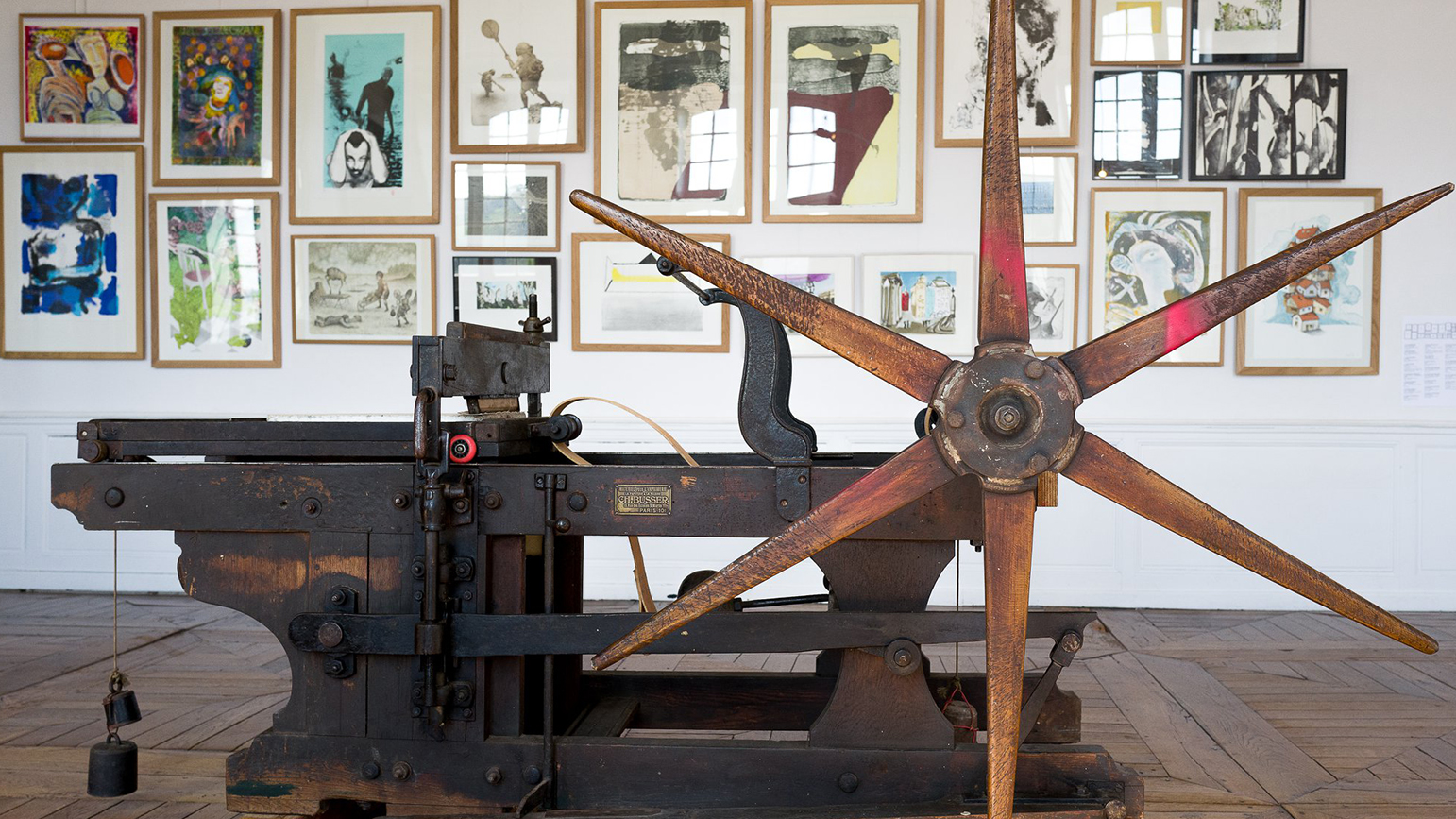 Photograph of a manual printing press with framed prints hung salon-style behind it.