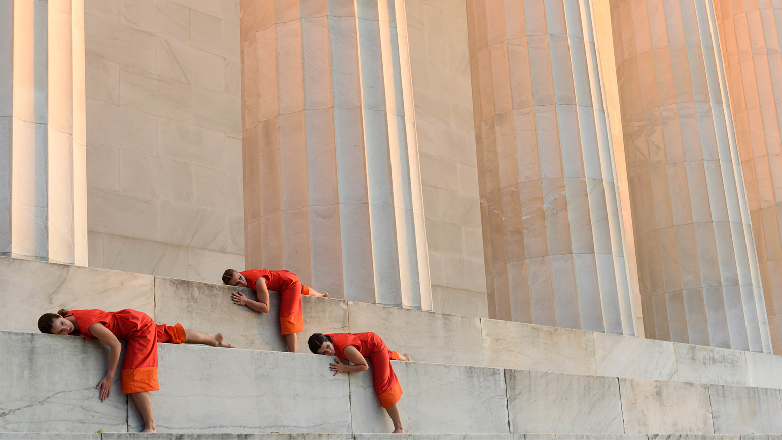 Women in orange jumpers lying on steps in front of a colonnade