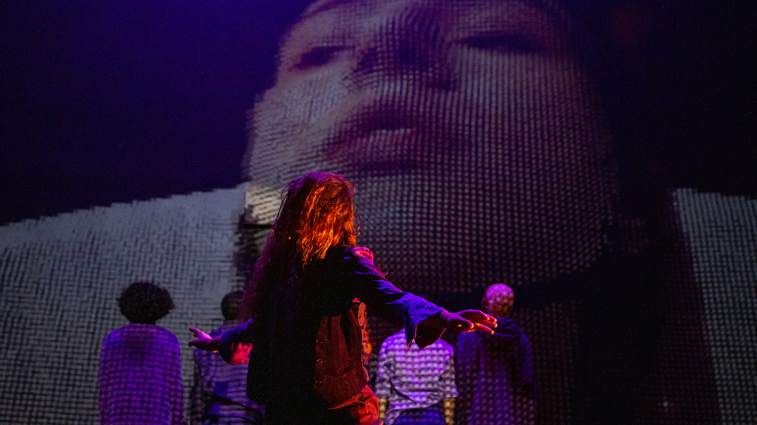 The stage saturated in purple. One dancer's hair covers her face with her arms out to the side, she stands in front of a close up pixelated projection of a white woman's face.