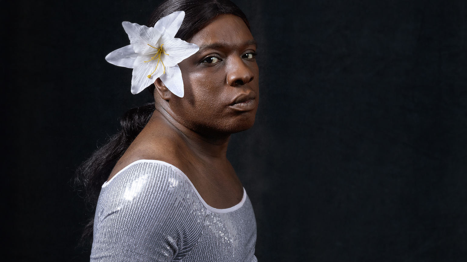 Bust of a person wearing a sparkly leotard with a white flower in their hair facing the camera