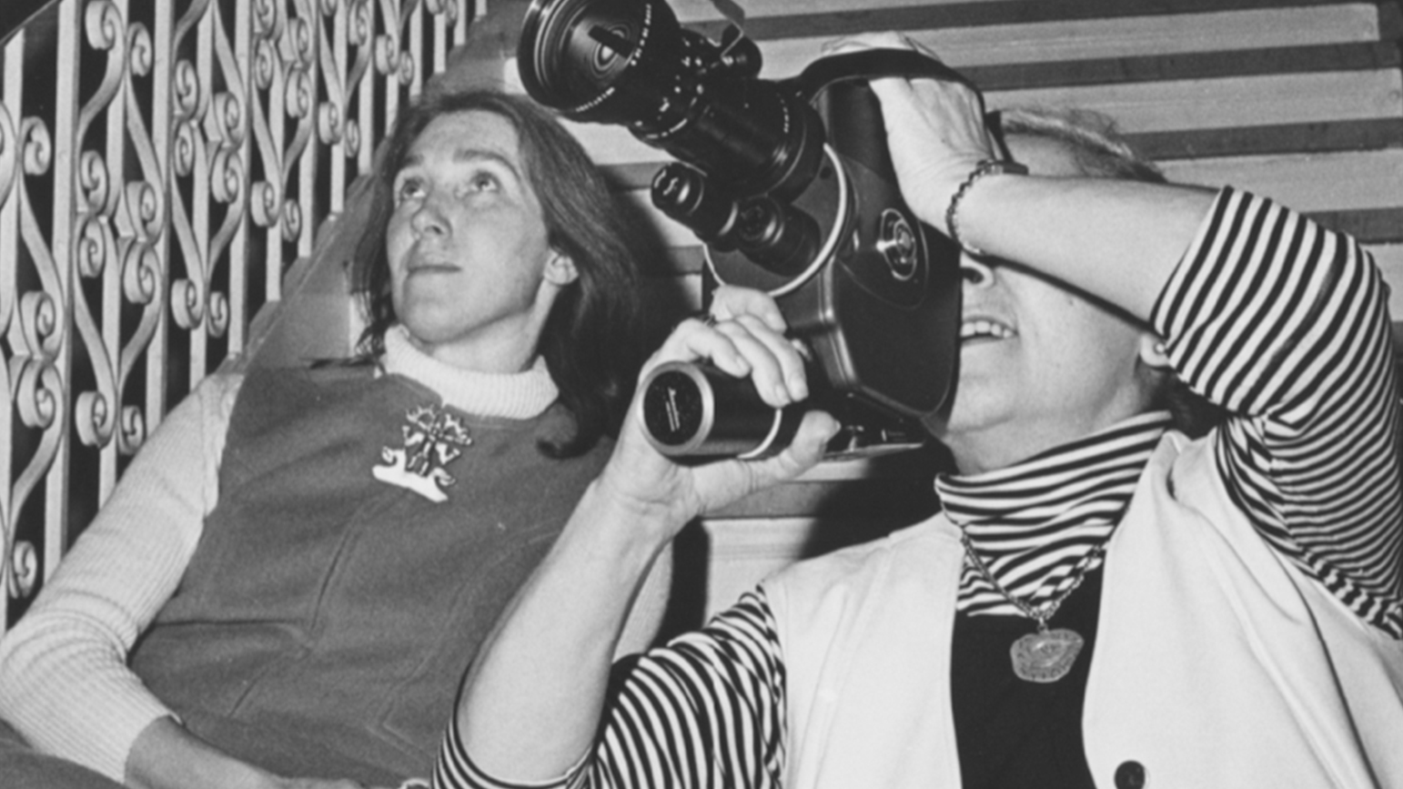 Black and white photograph of two women seated, one looking through a video camera