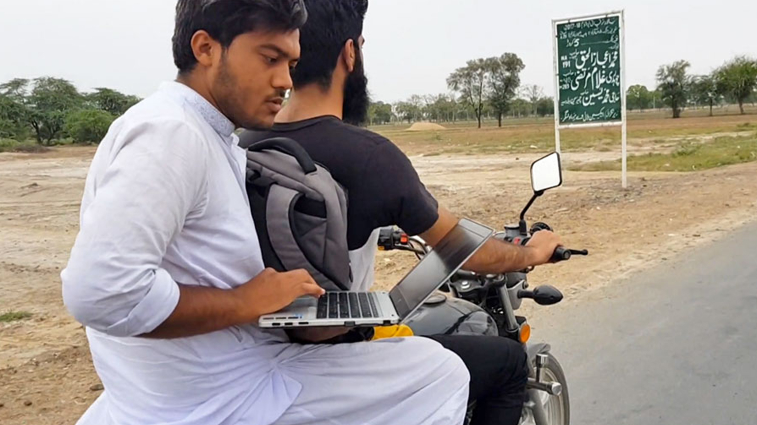 Photograph of two men on a motorcycle, one holding a laptop