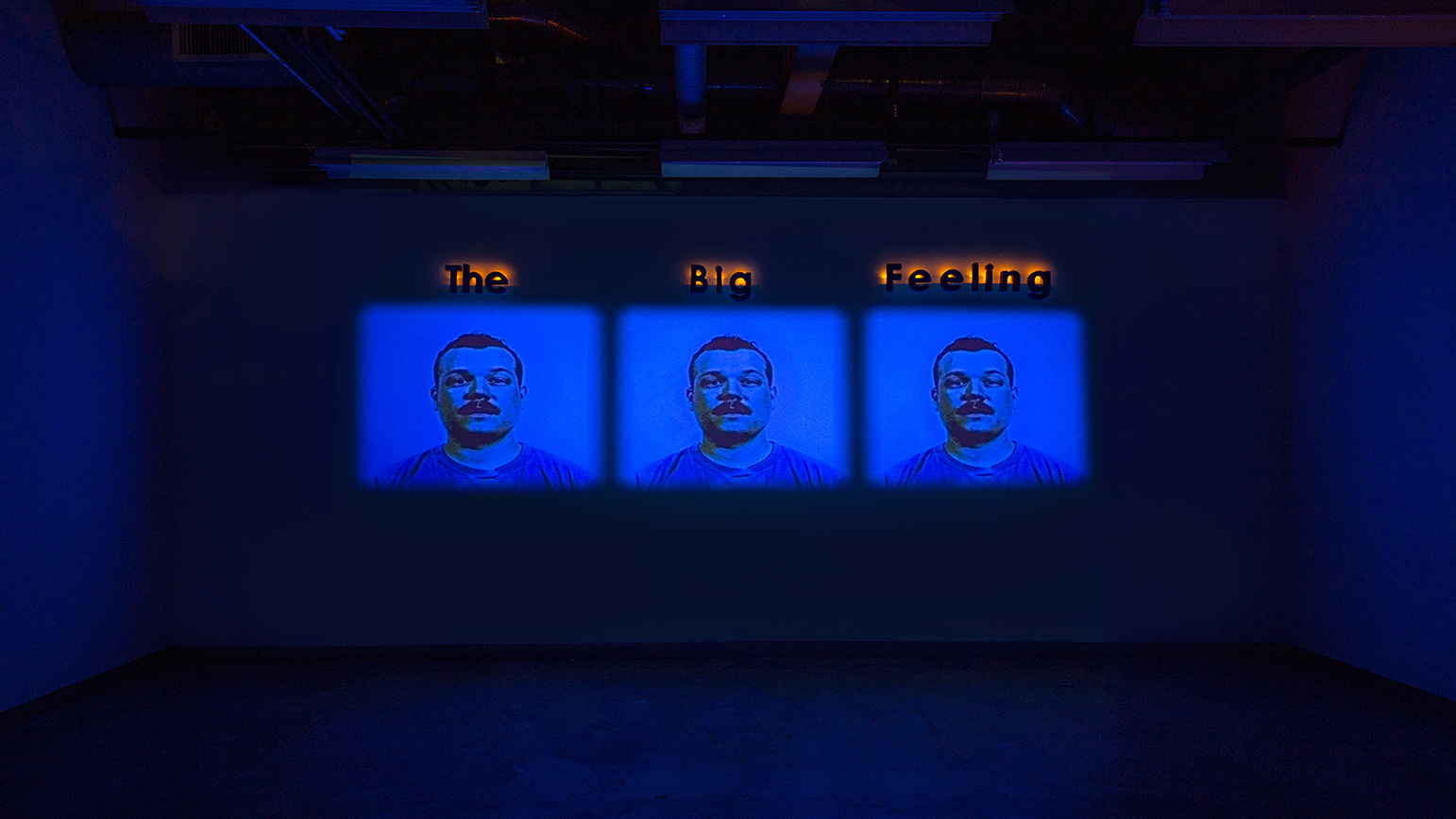 Three video projections showing the identical face with the works "The Big Feeling" above the projections