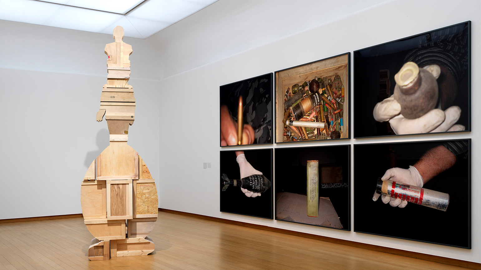 Installation view of an exhibition showing an abstract wood sculpture and photographs of instruments of war (bullets, bombs, etc.)