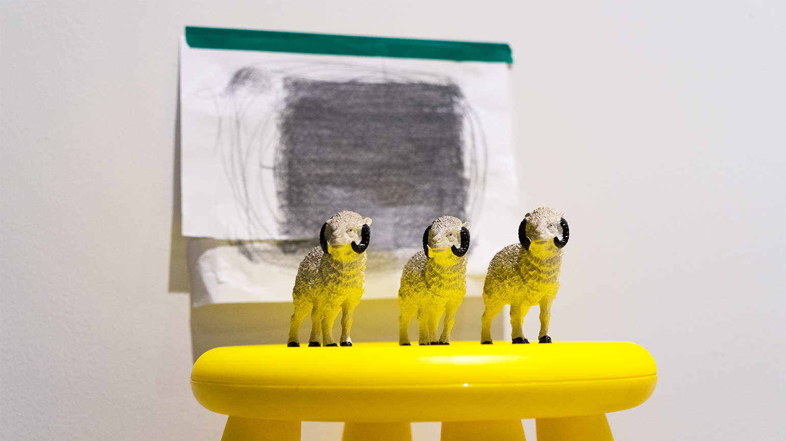 Three ram figurines on a stool with a drawing of a square taped on the wall behind