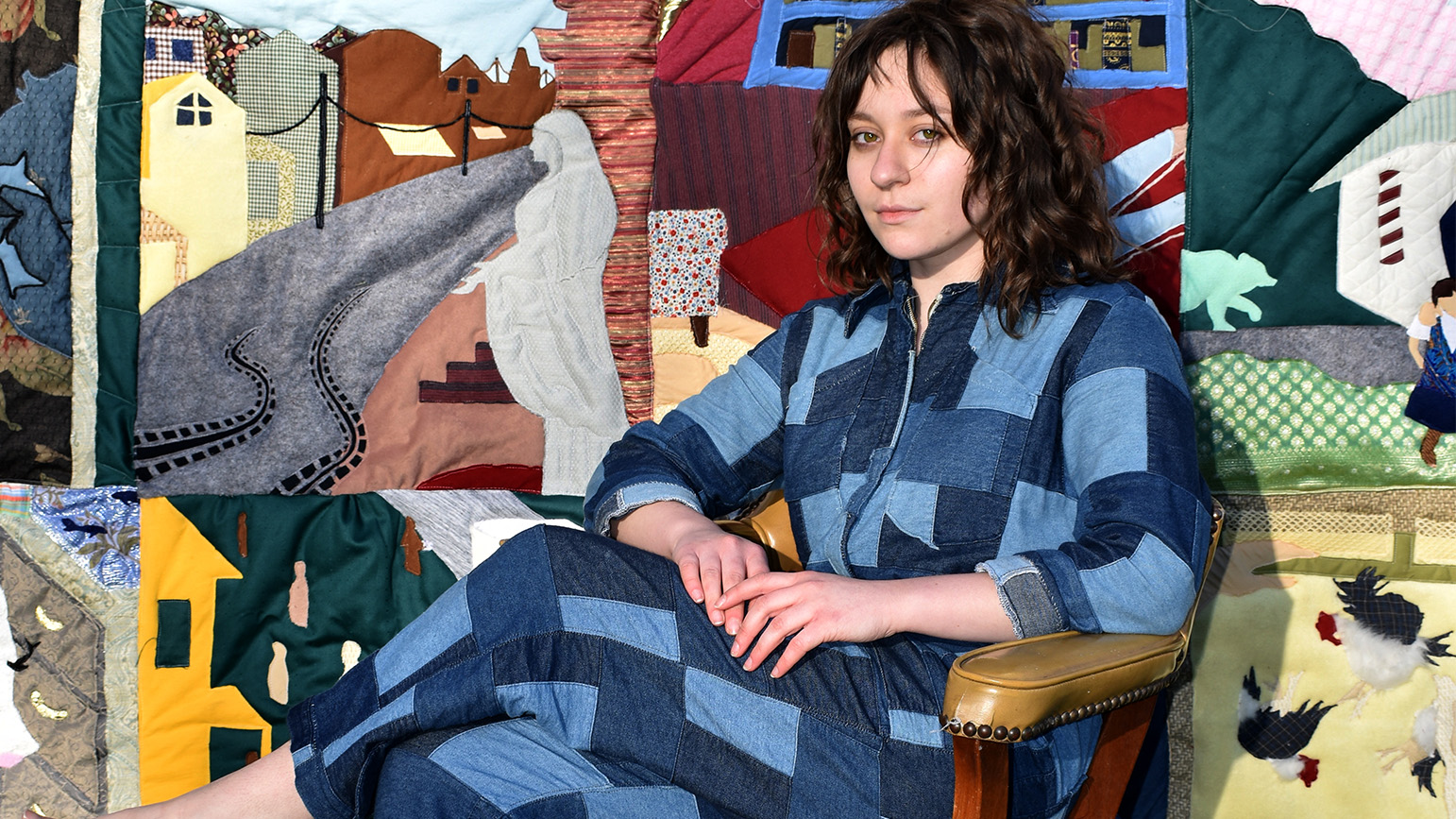 Photograph of Miranda Miller in a denim patchwork jumper in front of her family stories quilt