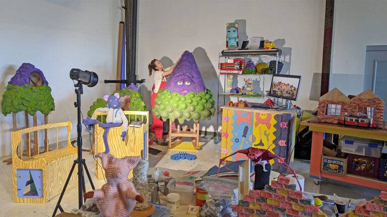 Studio filled with colorful, cartoon-like furniture and puppets. In the background, a woman is working on large purple sculpture with a cartoon face.