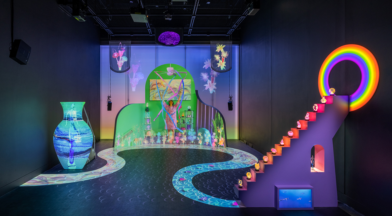 Installation image from Moulton's solo MoMA show with a large screen in the center, a vase-like shape with projection, a staircase leading to a rainbow halo, and a digital river.