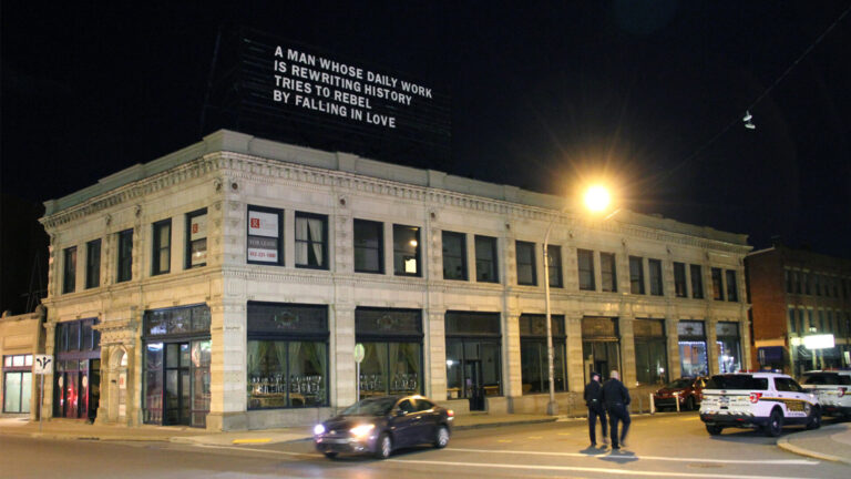 Two story building on a corner with a billboard on top that reads, in simple white text, "A Man Whose Daily Work is Rewriting History Tries to Rebel by Falling in Love."