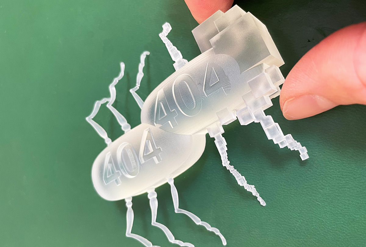 Close up of two fingers holding a plastic object of two bugs merged together with some parts more abstract. 404 is printed on both bugs.