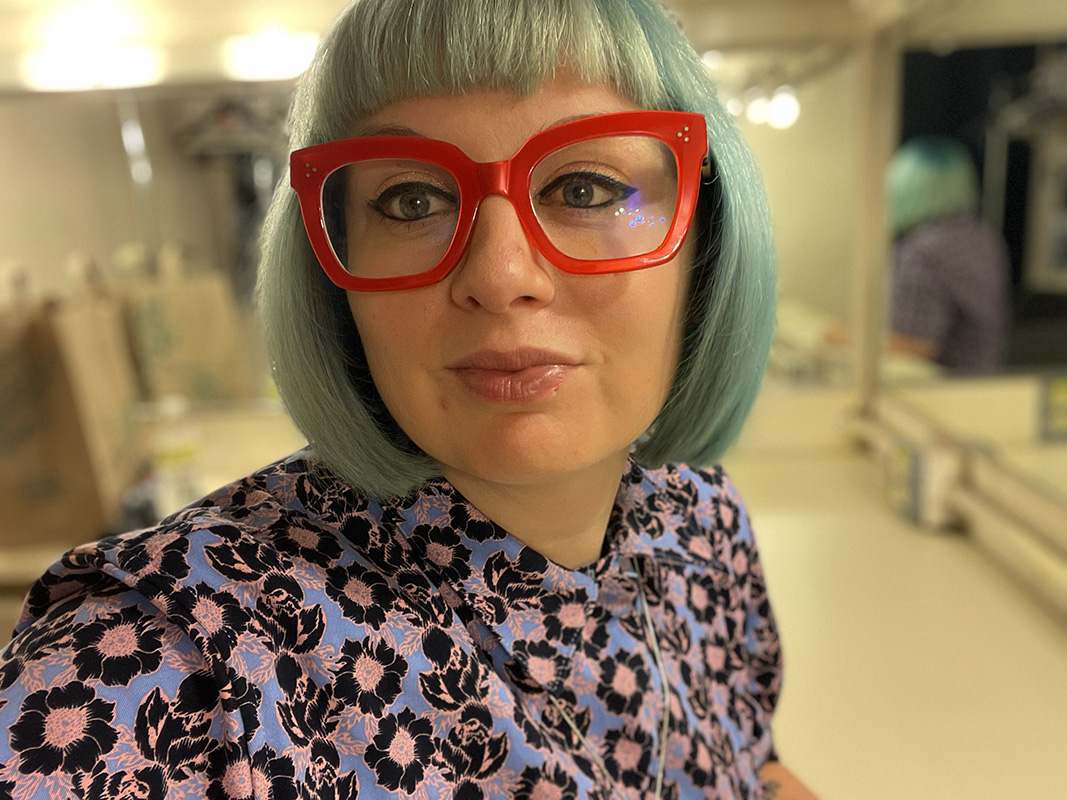 Portrait of Angela Waskho with short green hair and bright red glasses