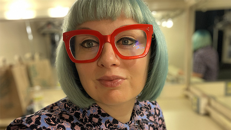 Portrait of Angela Waskho with short green hair and bright red glasses